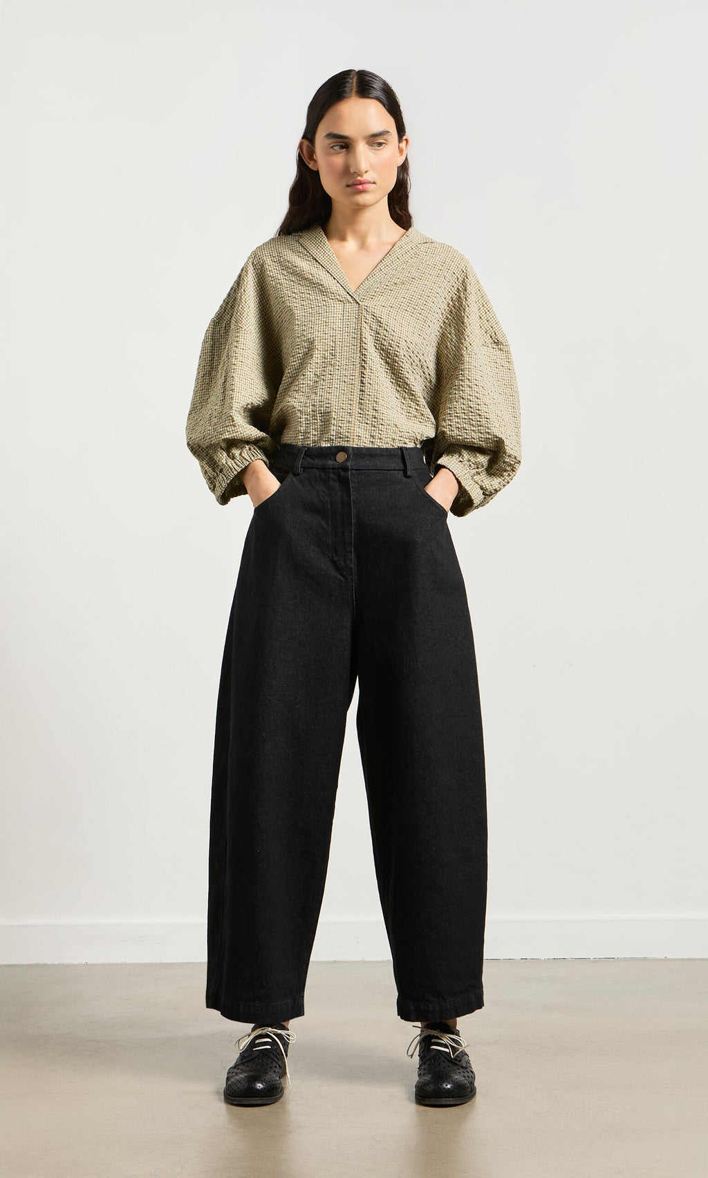Plümo- Woman wears Black Barrel Pants with hands in pockets, paired with a textured beige blouse against a plain background.