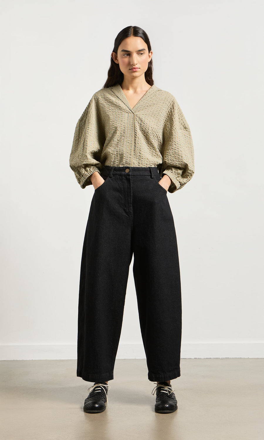 Plümo- Woman wears Black Barrel Pants with hands in pockets, paired with a textured beige blouse against a plain background.