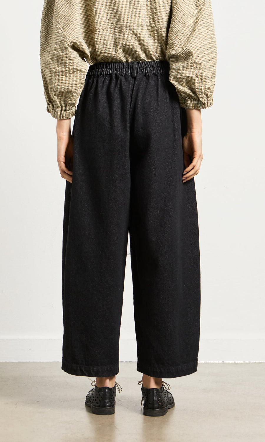 Plümo- Person stands on a light floor in relaxed silhouette, wearing Black Barrel Pants, black shoes, and beige long-sleeve top.