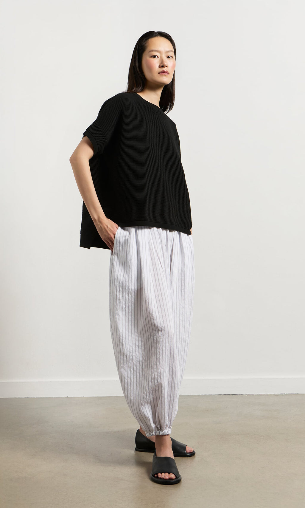 Plümo- Woman in a Jack Short Sleeve - Black top with white striped pants and sandals stands on a light floor by a plain wall.