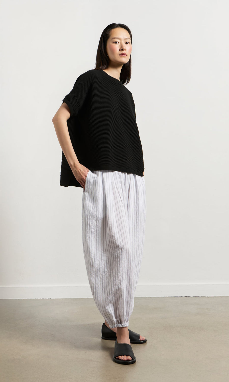 Plümo- Woman in a Jack Short Sleeve - Black top with white striped pants and sandals stands on a light floor by a plain wall.