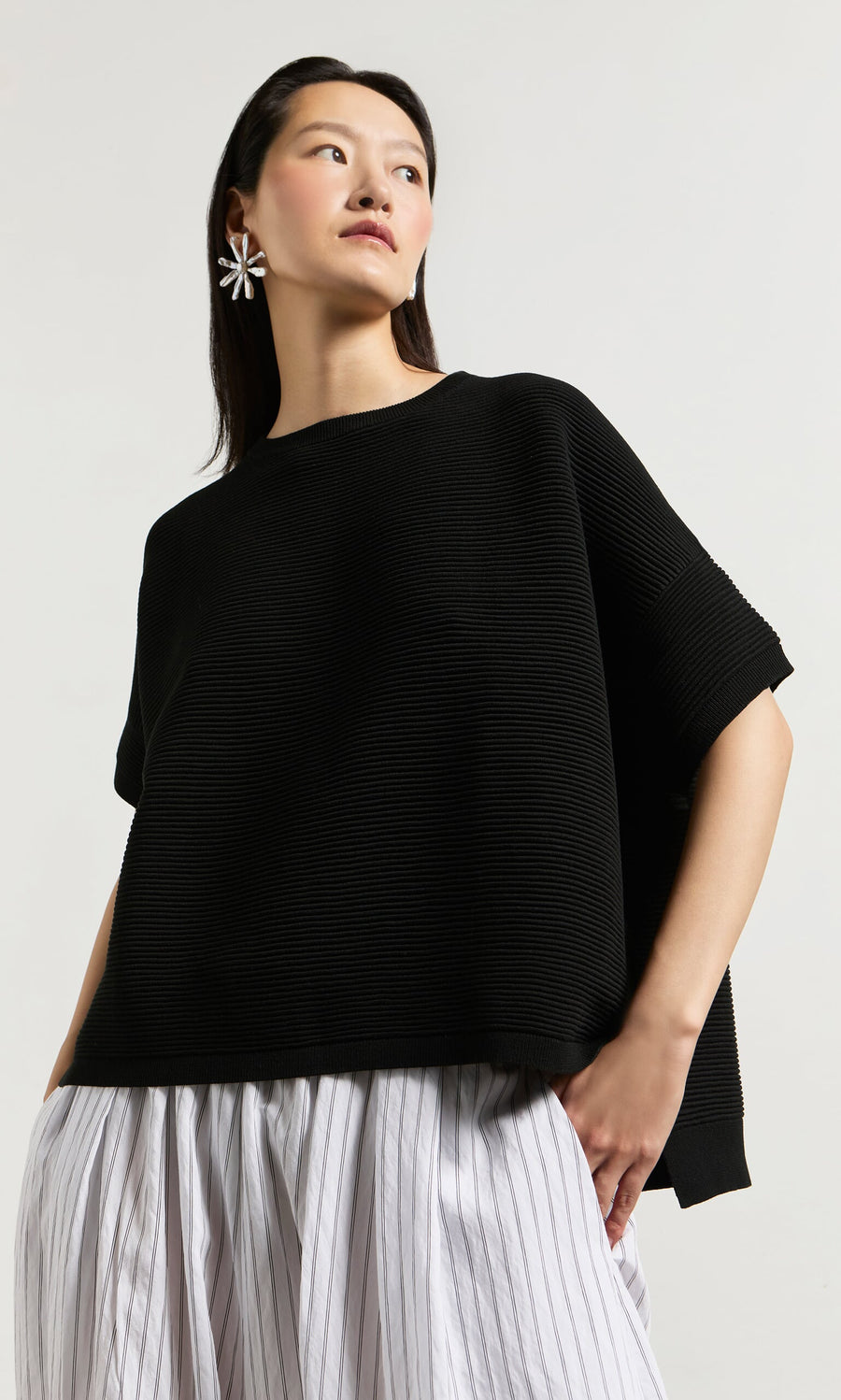 Plümo- Woman wears the Jack Short Sleeve - Black top with a white striped skirt and floral earrings, looking to the side.