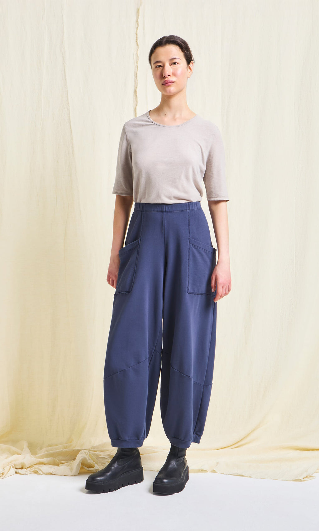 Plümo- A person stands against a cream backdrop, wearing a beige top, Blue Barrel Pants with large pockets, and black boots.