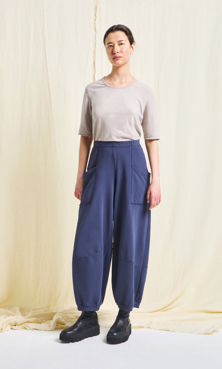 Plümo- A person stands against a cream backdrop, wearing a beige top, Blue Barrel Pants with large pockets, and black boots.