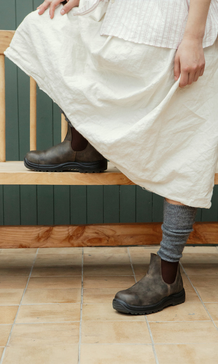 Plümo  Wearing Knitted Leg Warmers - Charcoal, person in white skirt and boots stands by a wooden bench on tiled floor.