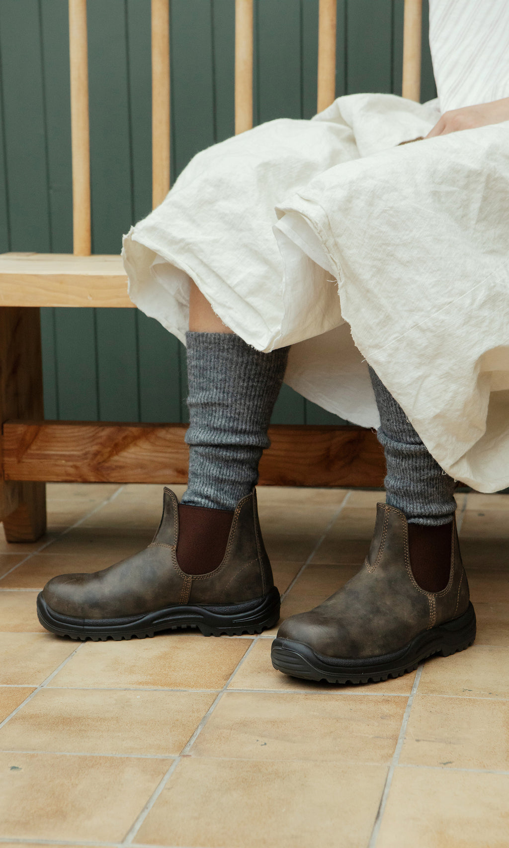 Plümo  Person in a white dress sits on a bench, wearing Knitted Leg Warmers - Charcoal and brown Chelsea boots on a tiled floor.