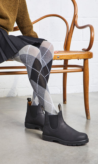 Plümo- Person in Blundstone Boots - Black, argyle tights, skirt, and brown sweater sits cross-legged on a wooden chair.