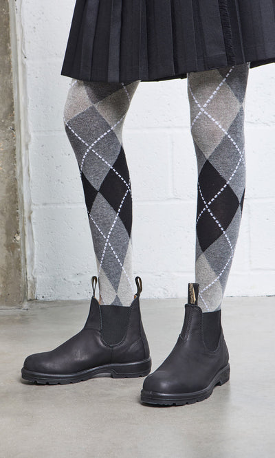 Plümo- Person in a black pleated skirt, gray argyle tights, and Blundstone Boots - Black on a concrete floor.