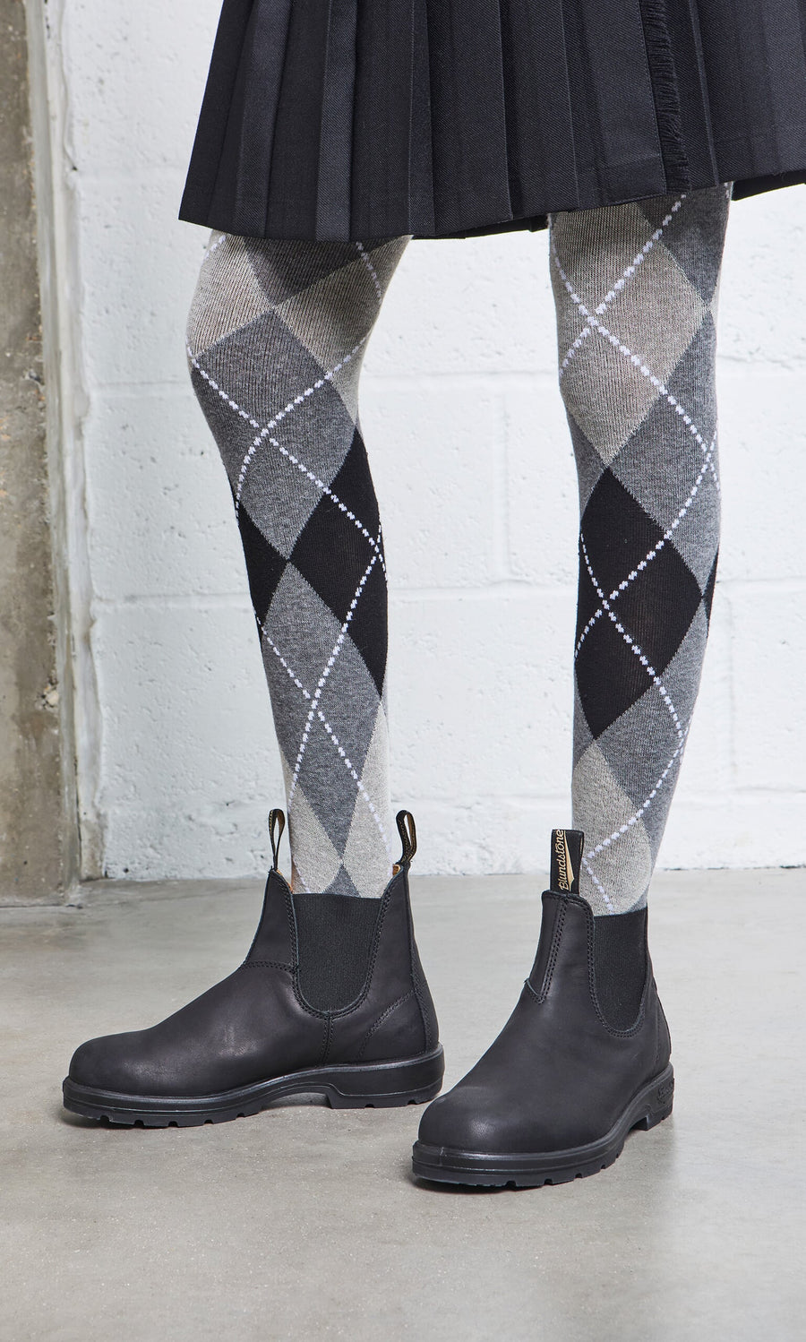Plümo- Person in a black pleated skirt, gray argyle tights, and Blundstone Boots - Black on a concrete floor.