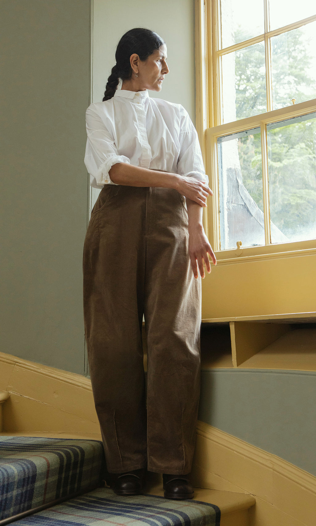 Plümo  Woman in a white shirt and Breda Pants stands on stairs, gazing out a sunny window.
