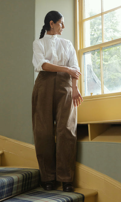 Plümo  Woman in a white shirt and Breda Pants stands on stairs, gazing out a sunny window.