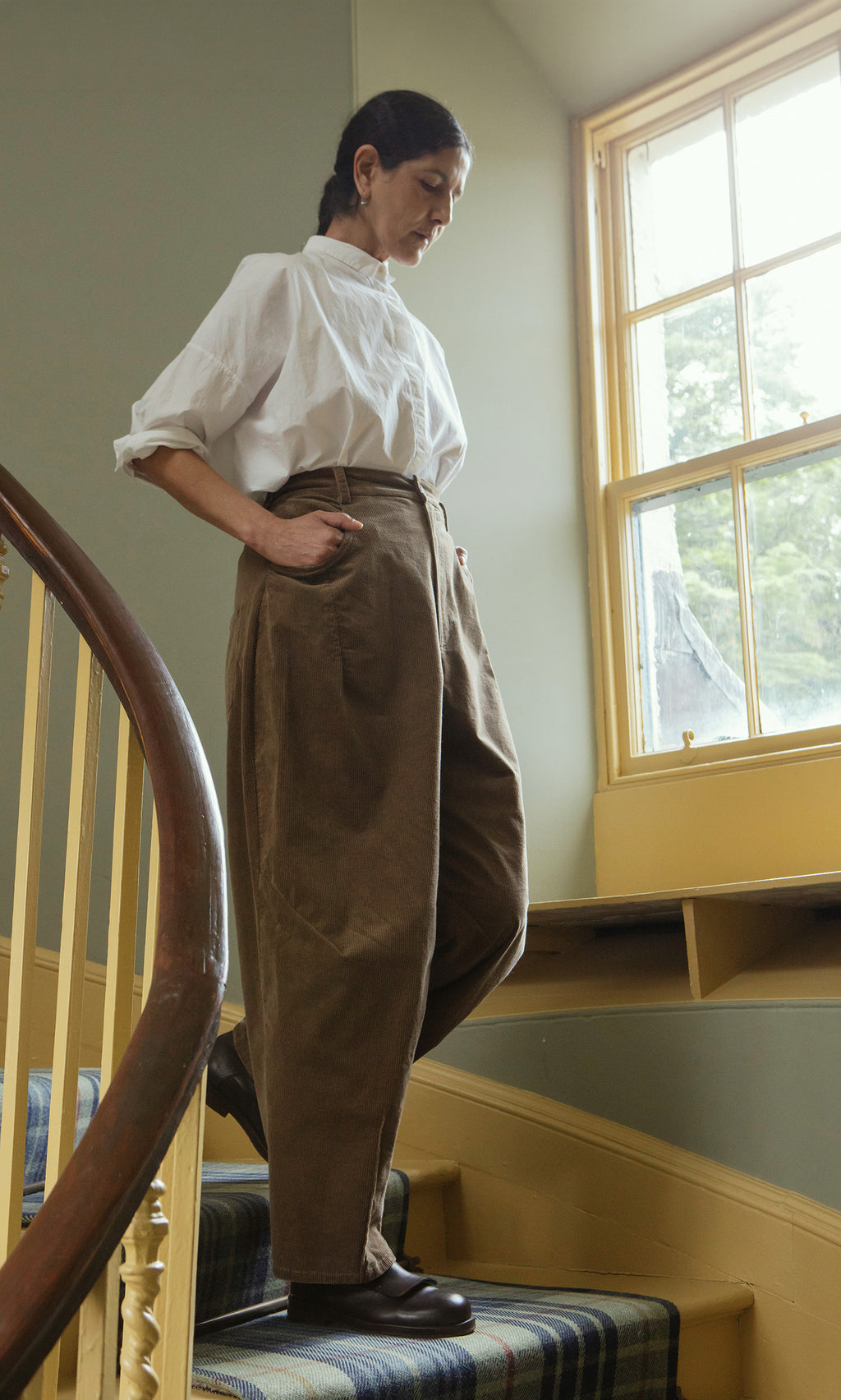 Plümo  Woman in white shirt and Breda Pants stands on a staircase by a window with natural light streaming in.