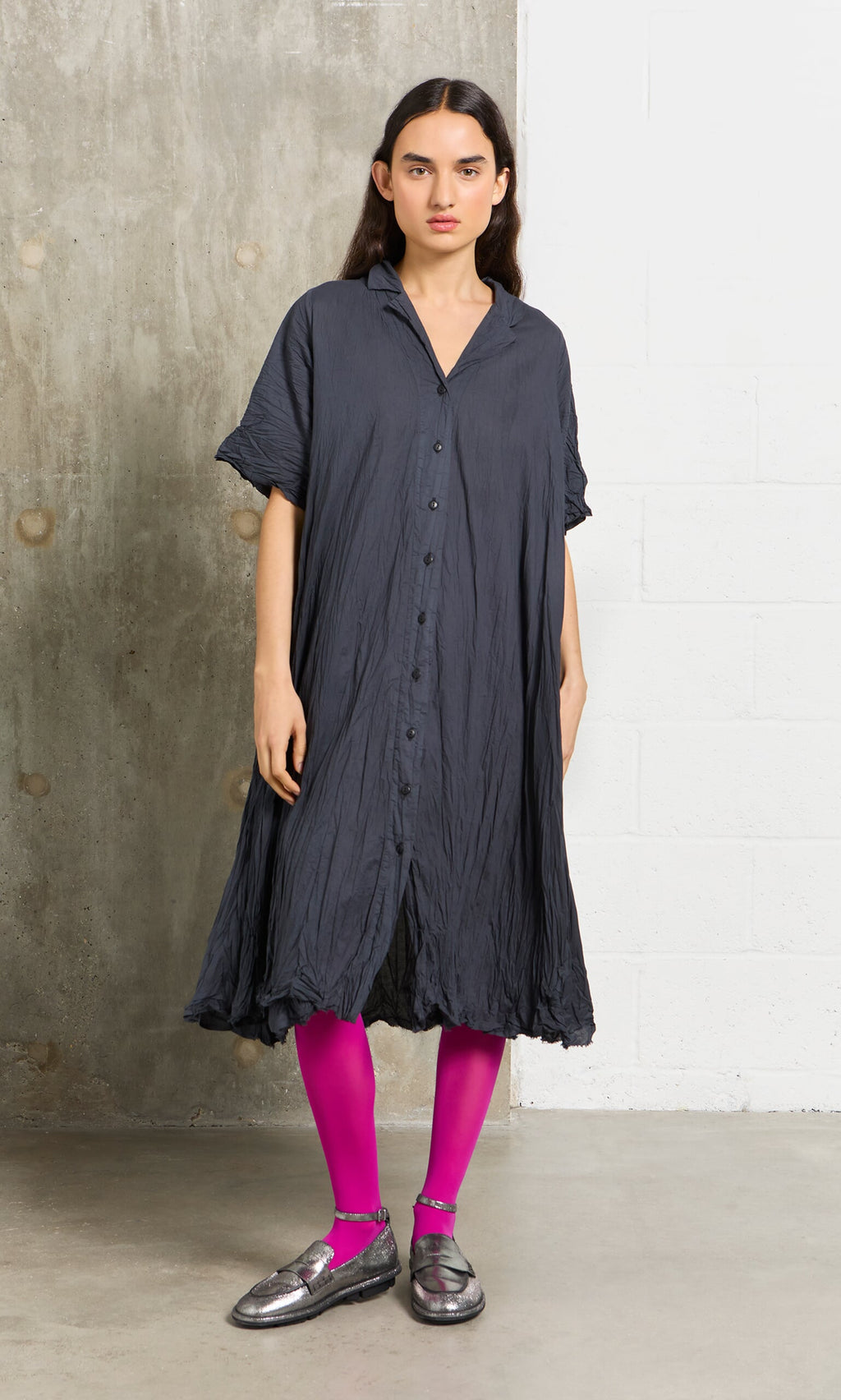 Plümo- A woman in a dark button-up dress wears Bright Magenta Tights, standing indoors against a gray wall.