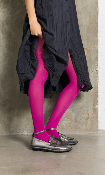 Plümo- Person in a black crinkled dress, Bright Magenta Tights, and silver shoes standing on a concrete floor.