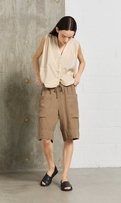 Plümo- A person in a beige sleeveless top and Brown Linen Shorts stands indoors against a concrete and white brick wall.