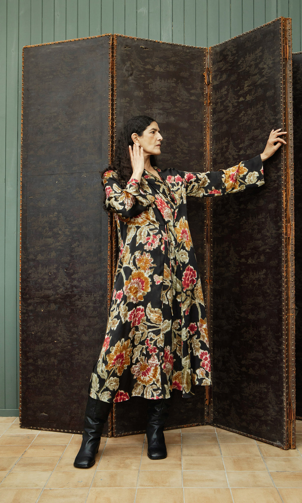 Plümo  Someone in a Brunn Dress and black boots poses by a dark folding screen, one hand raised to their ear.