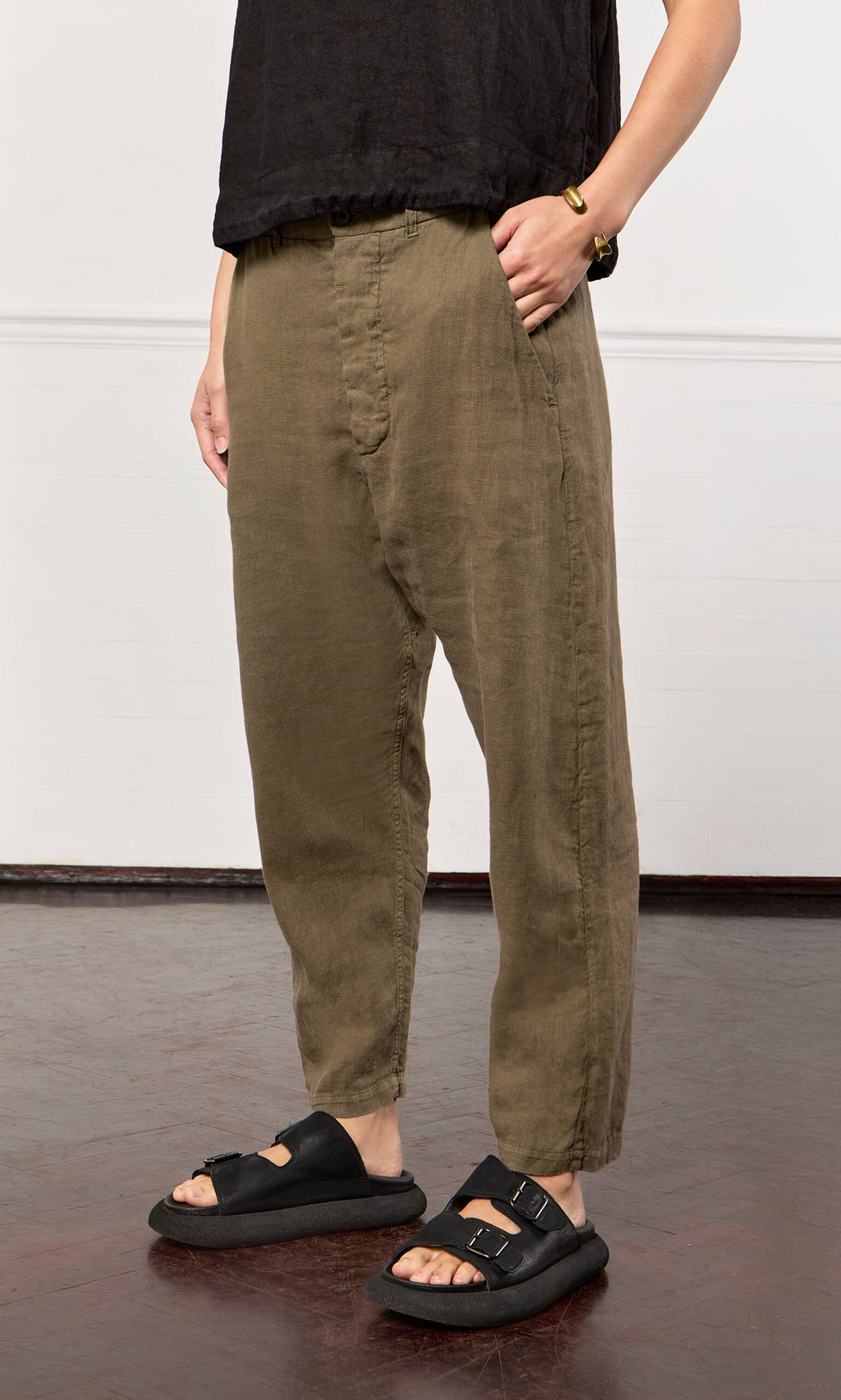 Plümo- Person wearing loose olive green Caper pants, a black top, black sandals, and a bracelet, standing indoors on a dark floor.