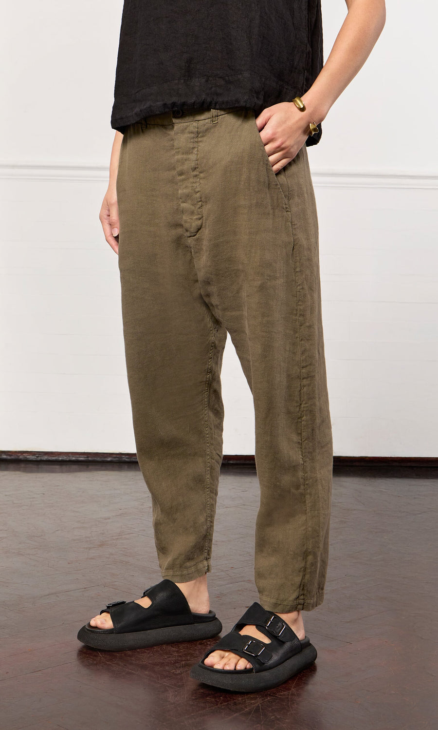 Plümo- Person wearing loose olive green Caper pants, a black top, black sandals, and a bracelet, standing indoors on a dark floor.