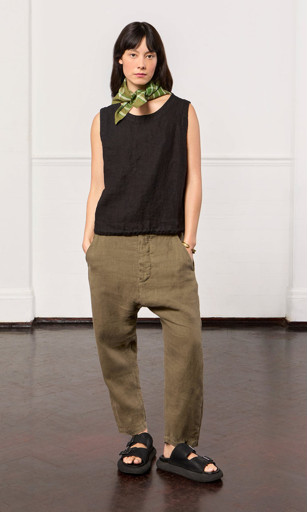 Plümo- Woman wearing Caper pants, a black sleeveless top, green neck scarf, and black sandals indoors.