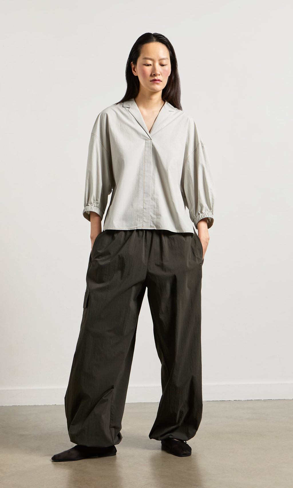 Plümo- Woman in light gray top and Cargo Pants stands against white wall, hands in pockets, wearing black shoes.