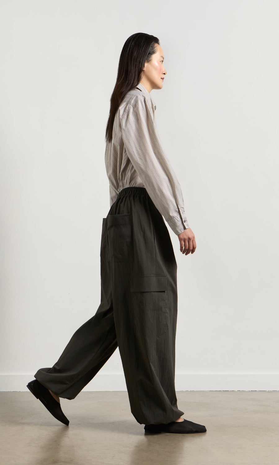 Plümo- Woman in cargo pants and a striped shirt walks indoors, facing right, against a plain white wall.