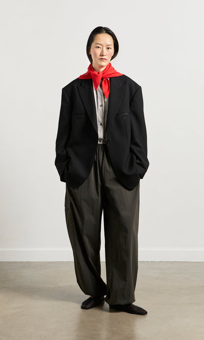 Plümo- Person in a black suit jacket, gray shirt, Cargo Pants with ankle bungee cords, and red scarf stands against a plain background.