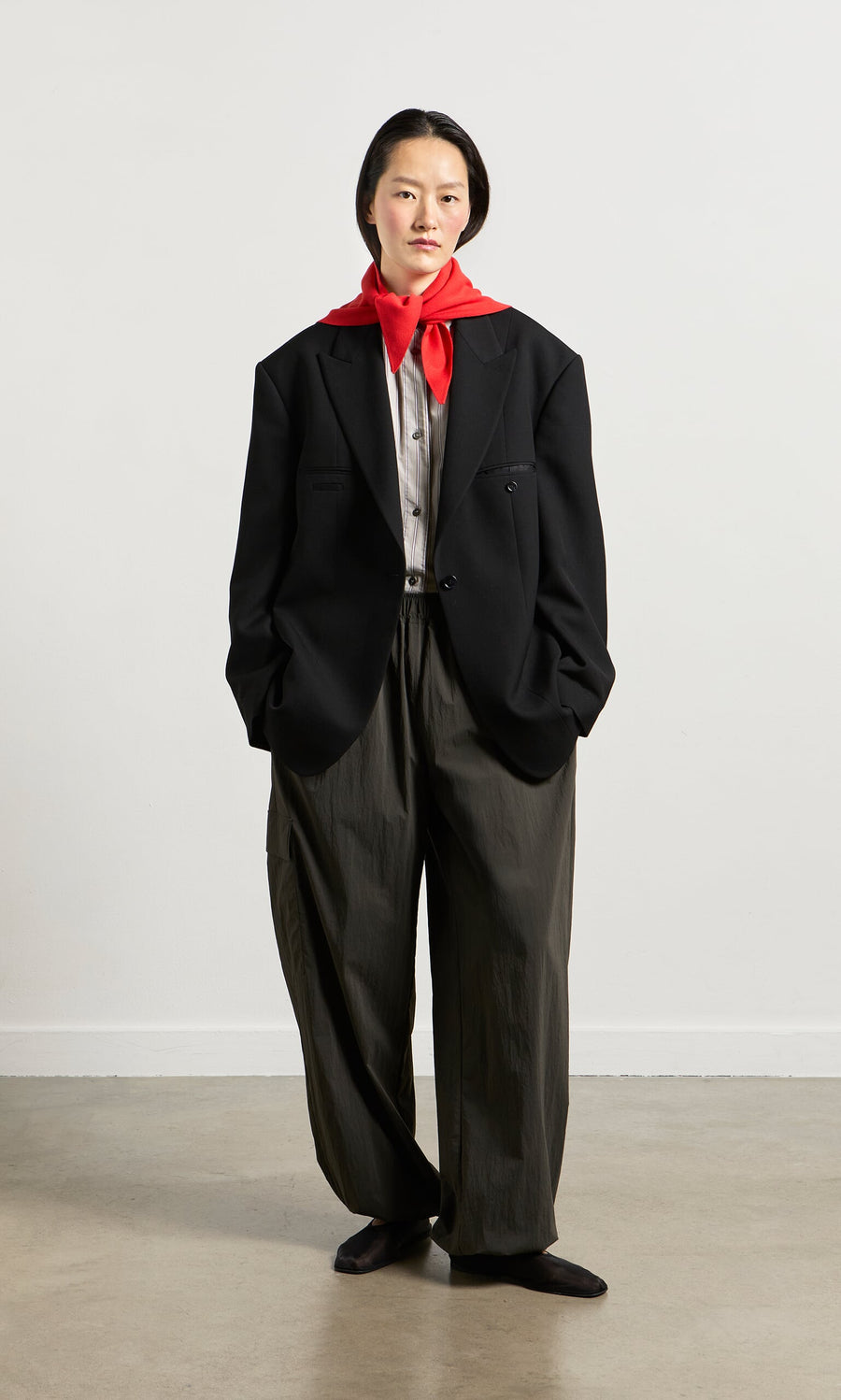 Plümo- Person in a black suit jacket, gray shirt, Cargo Pants with ankle bungee cords, and red scarf stands against a plain background.