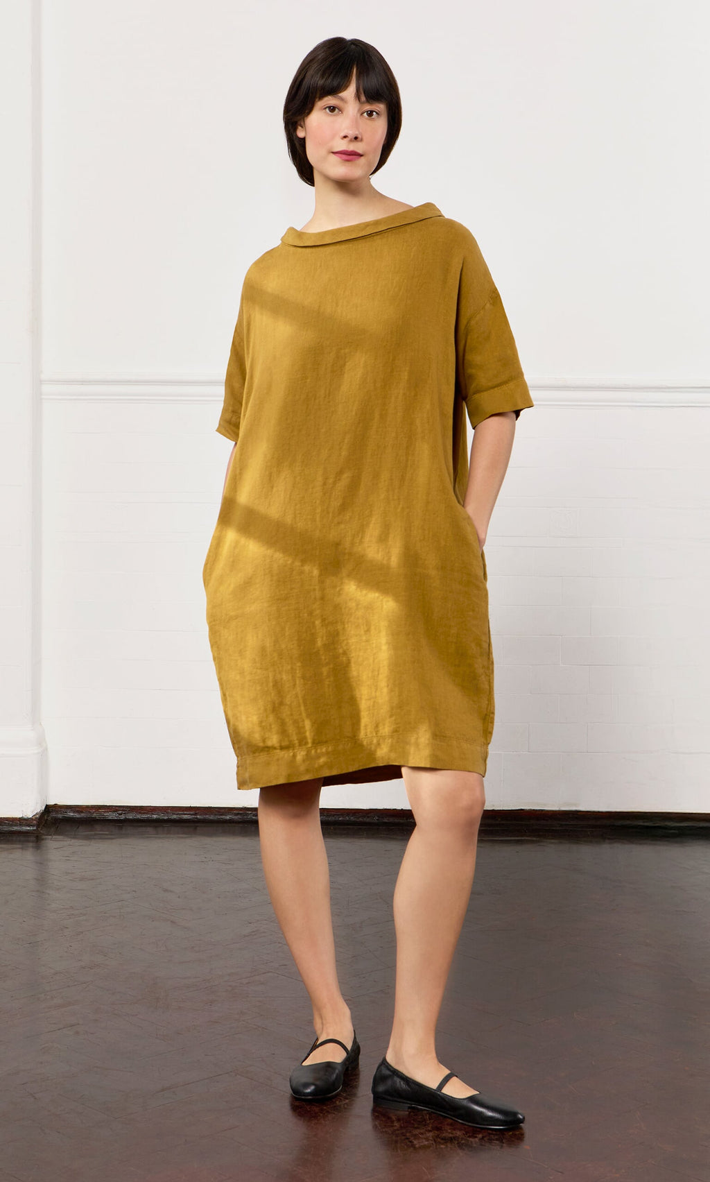 Plümo- A person stands indoors wearing the loose-fitting Carmone Dress with black flats, hands tucked in its pockets.