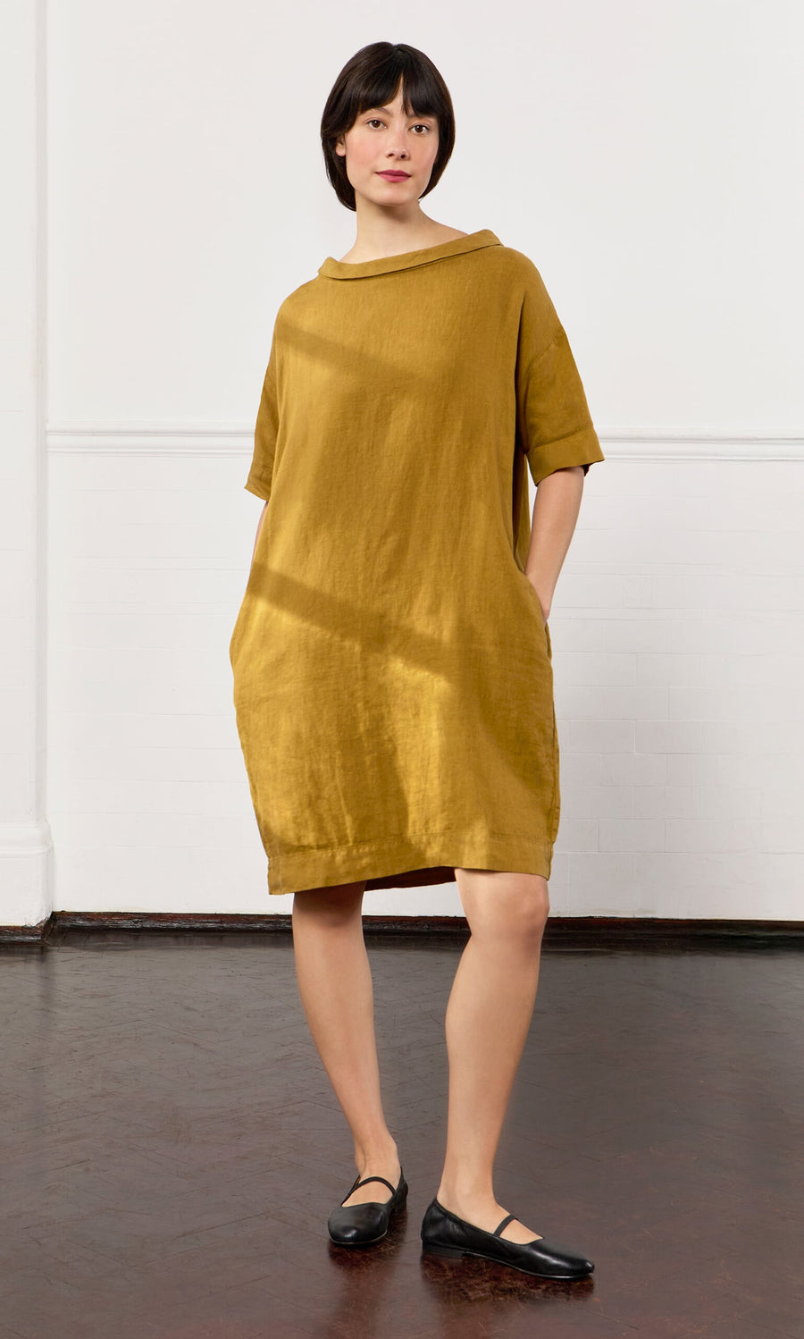 Plümo- A person stands indoors wearing the loose-fitting Carmone Dress with black flats, hands tucked in its pockets.