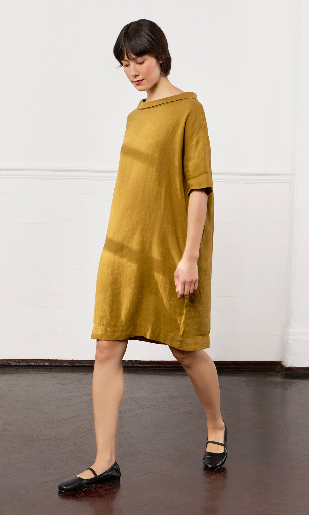Plümo- Woman in a loose mustard yellow Carmone Dress and black shoes walking indoors on a dark wood floor.