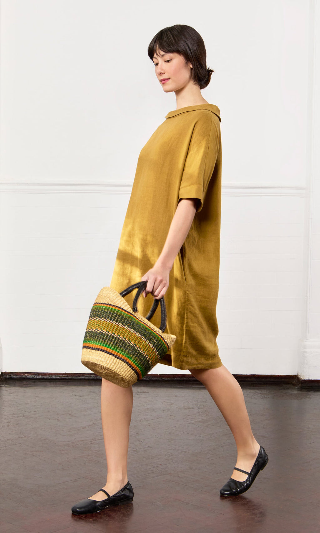 Plümo- Woman in the Carmone Dress holds a woven handbag, walking left on a dark floor against a white wall.