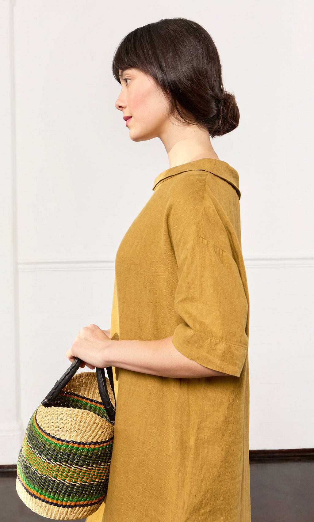 Plümo- Woman in the Carmone Dress, a mustard yellow linen piece, holding a basket in profile on a plain white background.