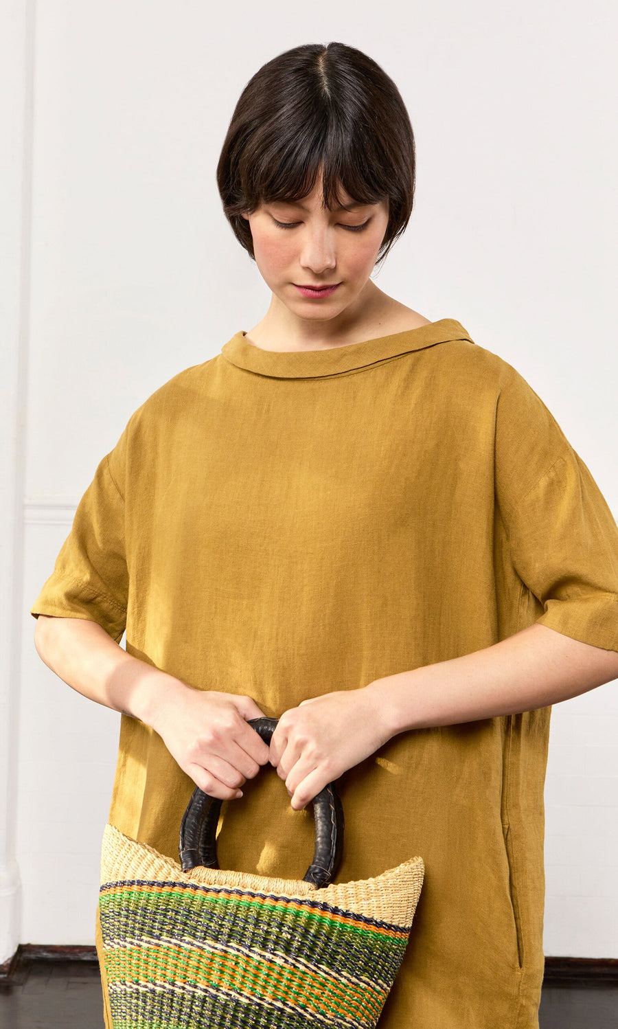 Plümo- Woman in the loose Carmone Dress in mustard yellow linen, holding a woven bag with dark handles against a plain background.