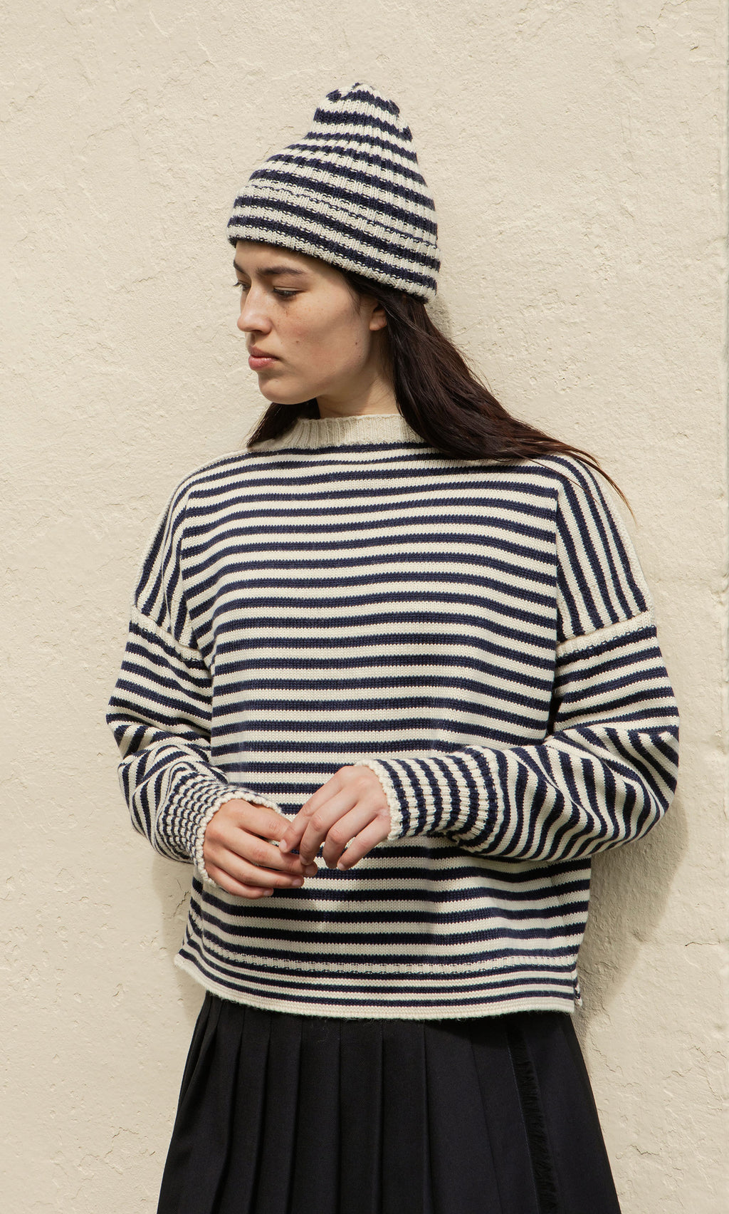 Plümo  Model in the Carnac Jumper stands by a textured beige wall, gazing sideways.