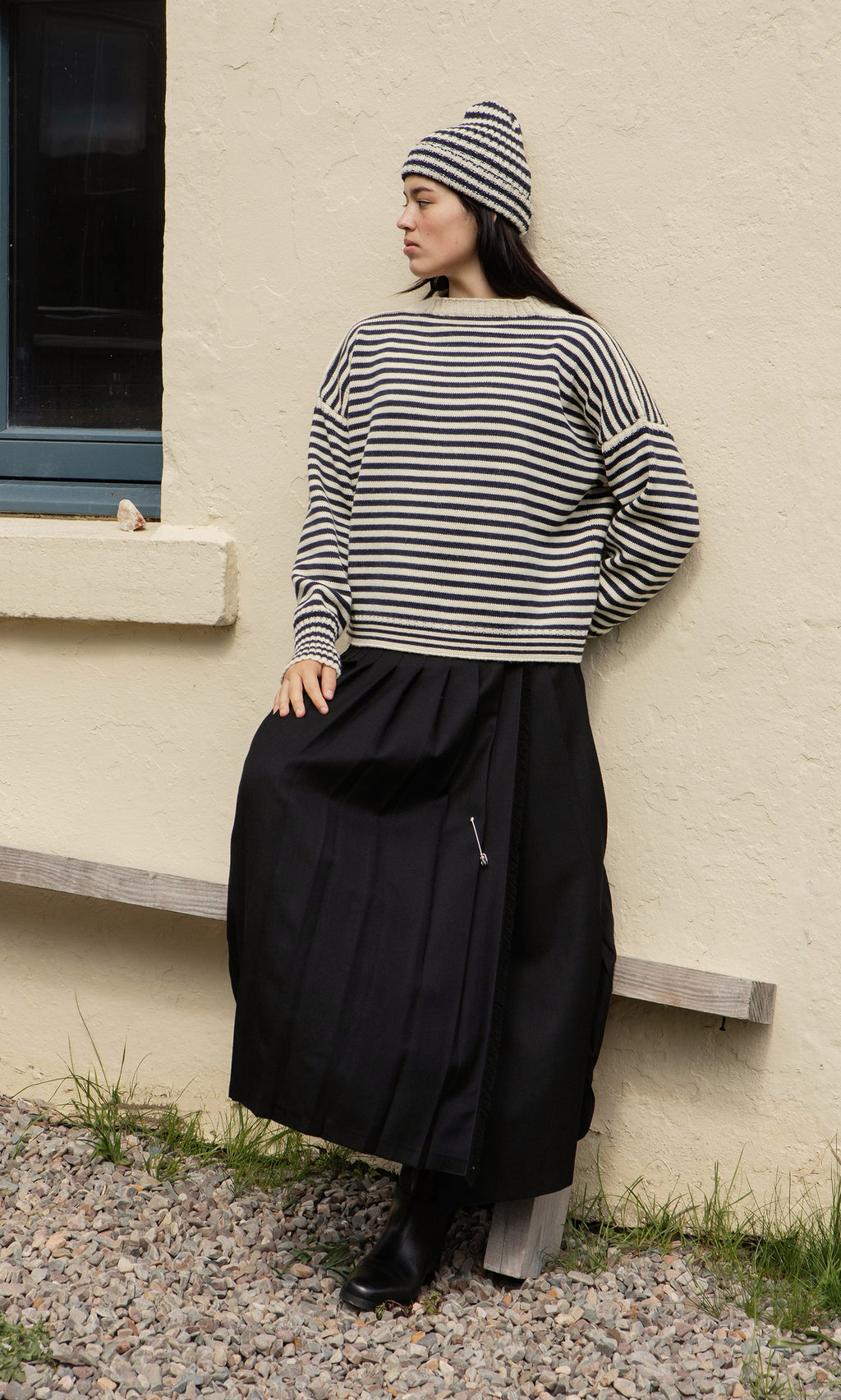 Plümo  Woman in Carnac Jumper, hat, long black skirt, and boots leans against windowed wall on wooden bench.