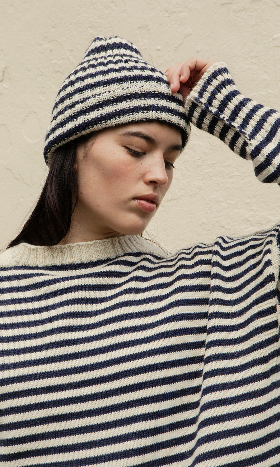 Plümo  Woman in a Carnac Jumper and hat stands against a textured wall, eyes gently closed.