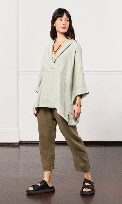 Plümo- Woman in Carvey Tunic, olive pants, black sandals, layered necklace, in a minimal indoor space.