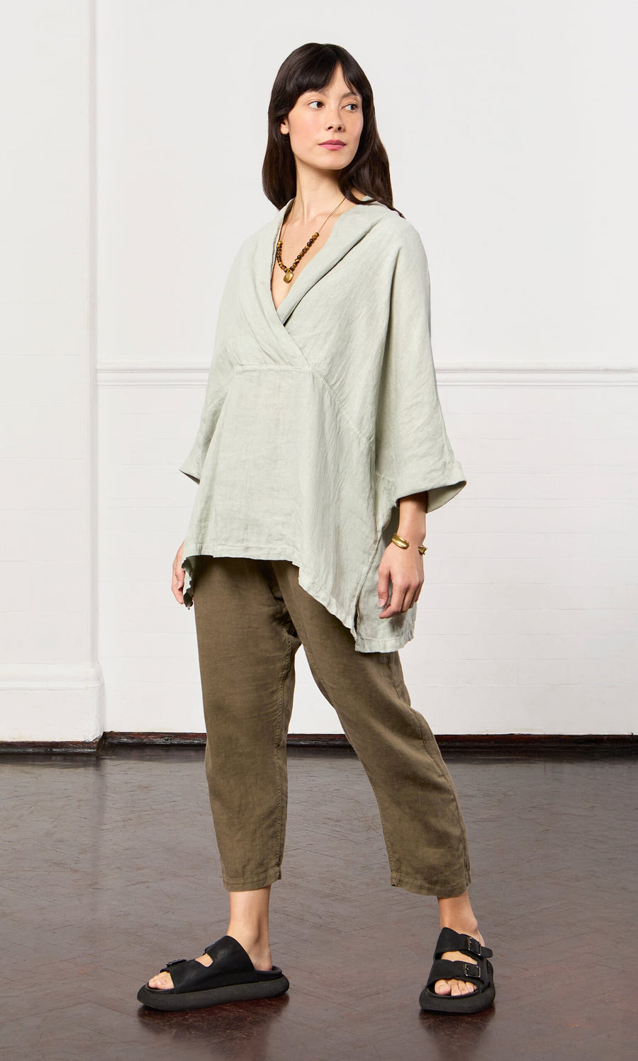 Plümo- Woman in Carvey Tunic, olive pants, black sandals, layered necklace, in a minimal indoor space.
