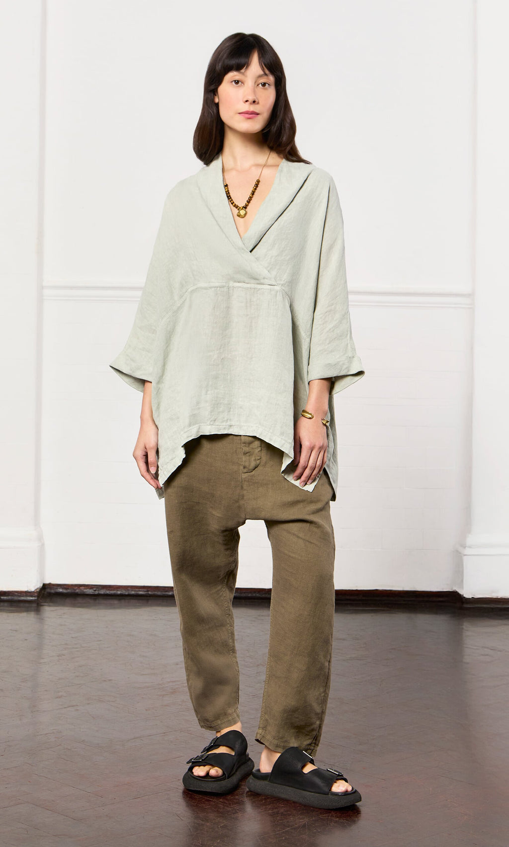 Plümo- Woman in a Carvey Tunic with cropped sleeves, olive pants, black sandals, and gold jewelry on a wooden floor.