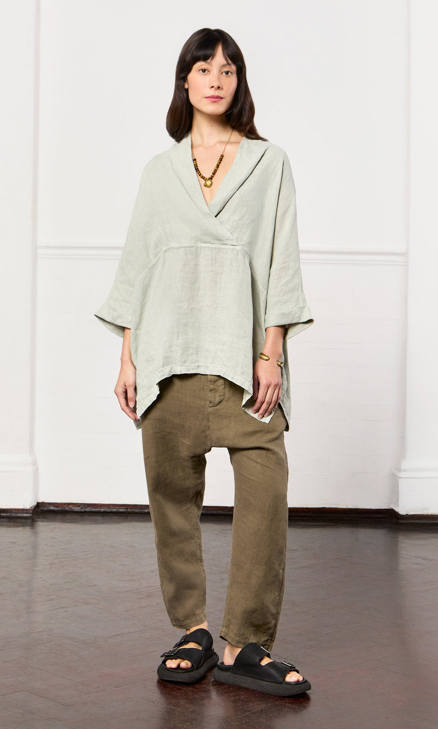 Plümo- Woman in a Carvey Tunic with cropped sleeves, olive pants, black sandals, and gold jewelry on a wooden floor.