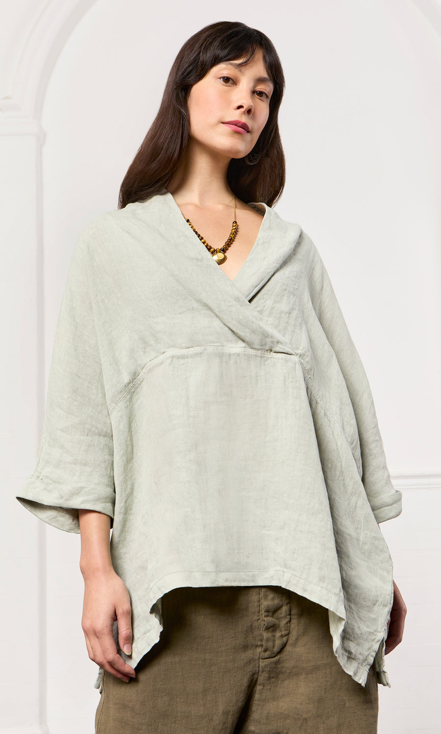 Plümo- Woman wearing the Carvey Tunic with cropped sleeves and olive pants, accessorized with a gold necklace indoors.