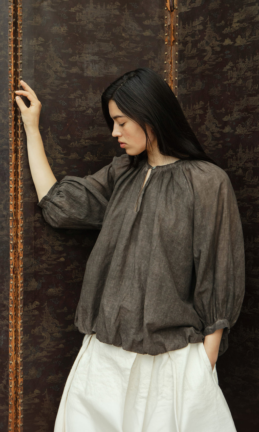Plümo  Woman in a loose Cinnamon Blouse and white pants stands pensively by a patterned, dark brown screen.