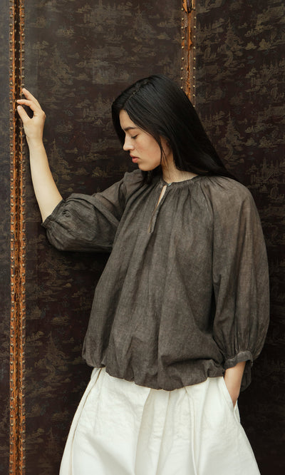 Plümo  Woman in a loose Cinnamon Blouse and white pants stands pensively by a patterned, dark brown screen.