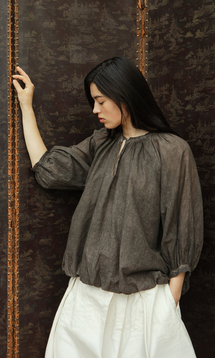 Plümo  Woman in a loose Cinnamon Blouse and white pants stands pensively by a patterned, dark brown screen.