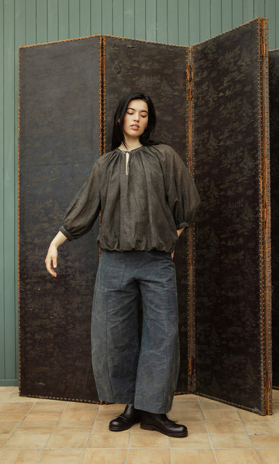 Plümo  Person in a loose Cinnamon Blouse and wide blue pants stands by a brown folding screen on a tiled floor.