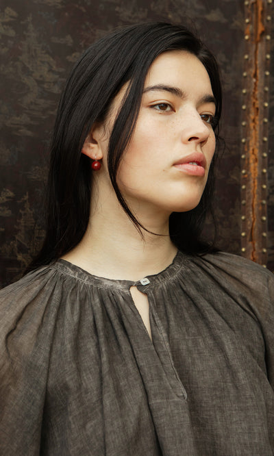 Plümo  Woman with long dark hair wears the Cinnamon Blouse and red earrings, standing against a textured background.