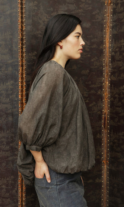 Plümo  Woman stands sideways in Cinnamon Blouse, hands in pockets, against a dark patterned background, looking ahead.