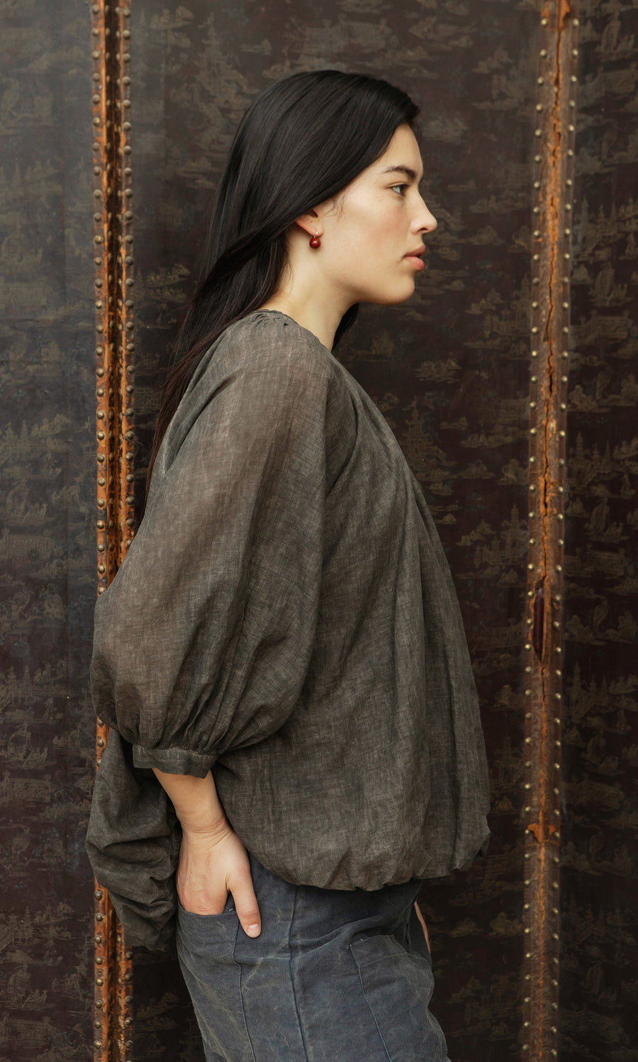 Plümo  Woman stands sideways in Cinnamon Blouse, hands in pockets, against a dark patterned background, looking ahead.
