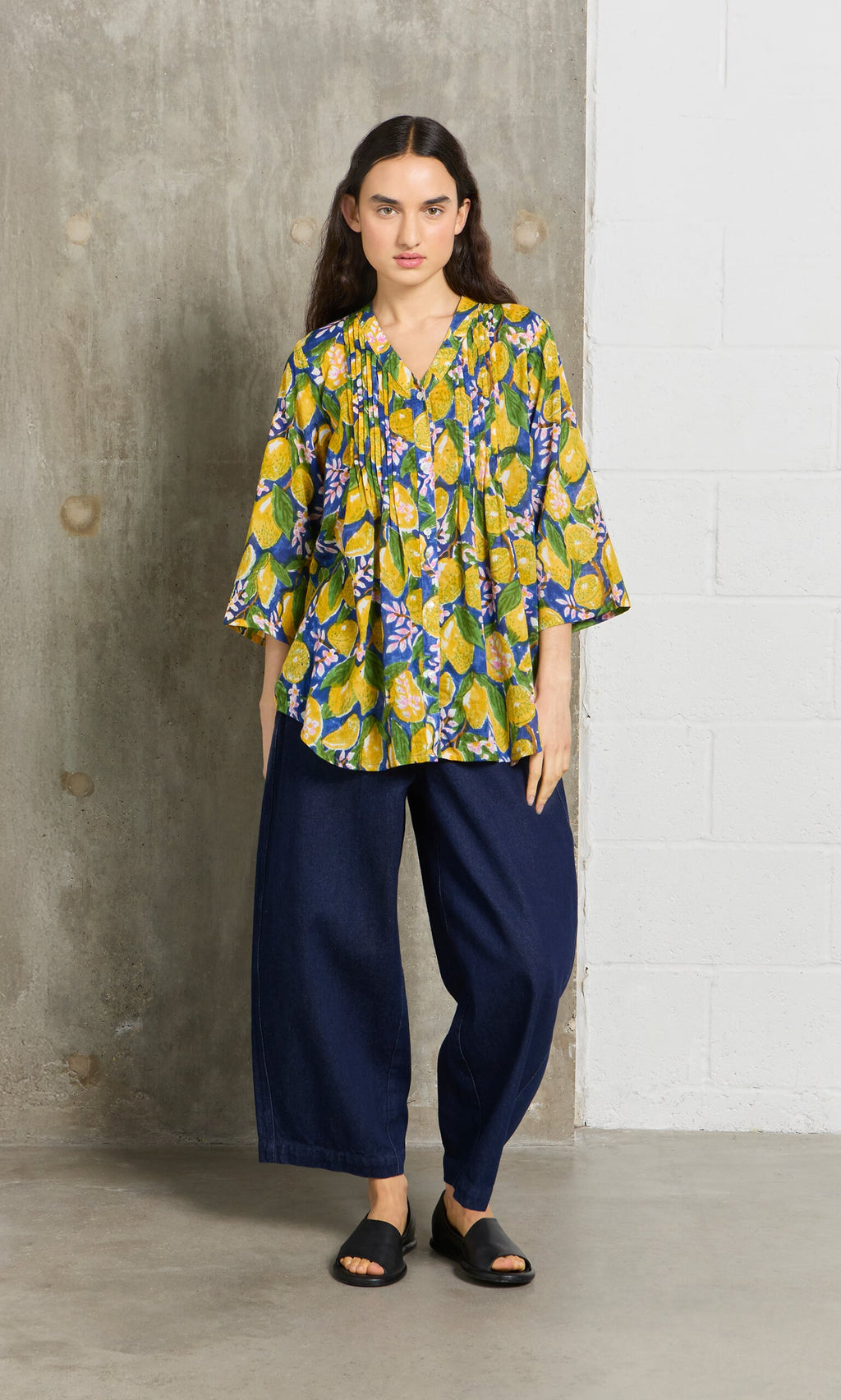 Plümo- Woman in a hand block-printed Citrus Shirt, wide-leg dark blue pants, and black sandals stands by a concrete wall.