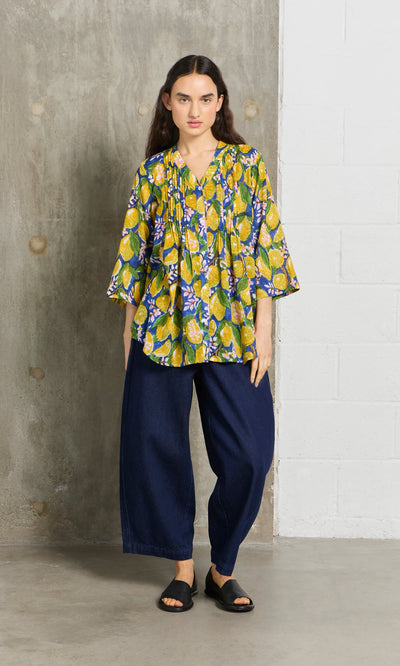 Plümo- Woman in a hand block-printed Citrus Shirt, wide-leg dark blue pants, and black sandals stands by a concrete wall.